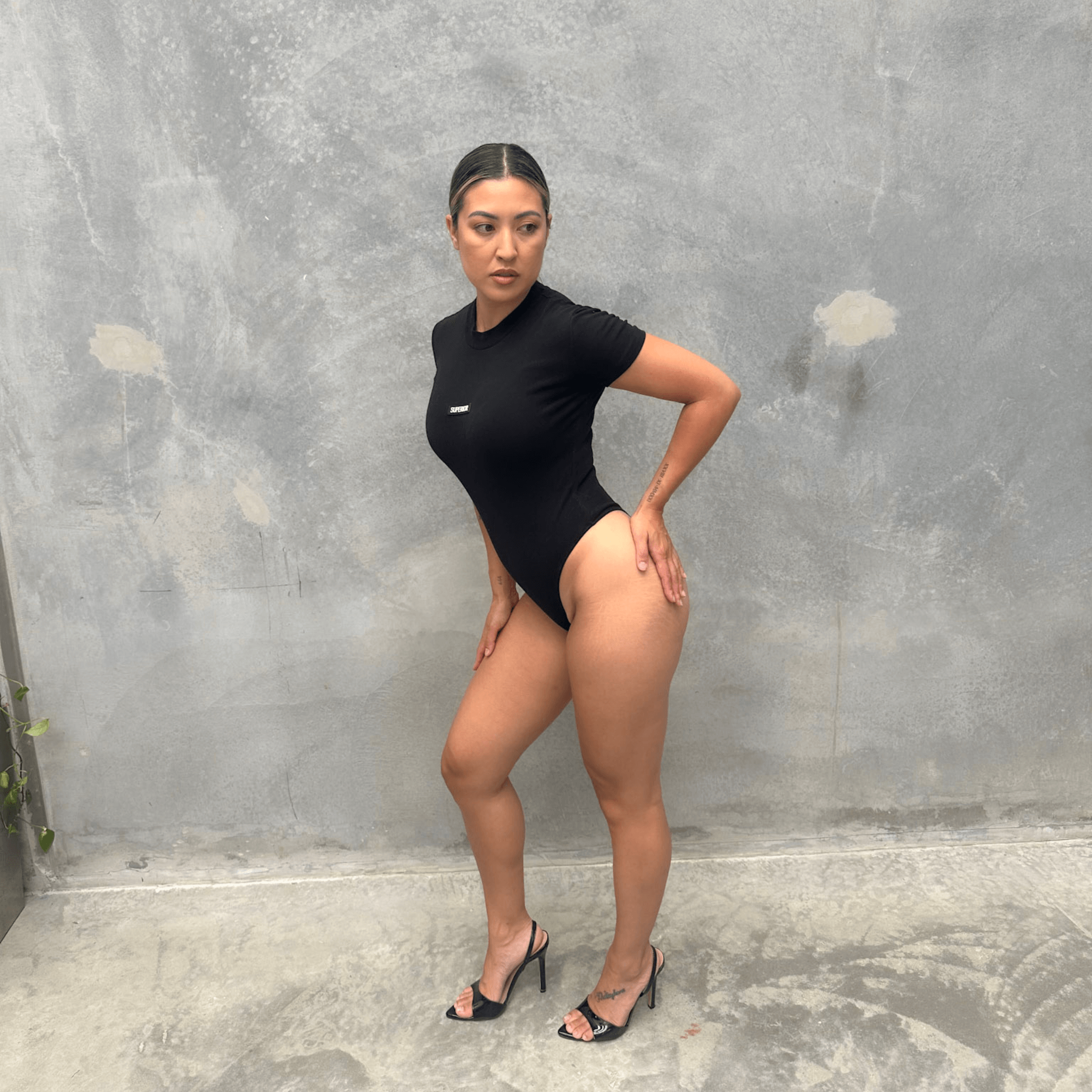 Woman in a black short sleeve bodysuit against a grey concrete wall posing from the side
