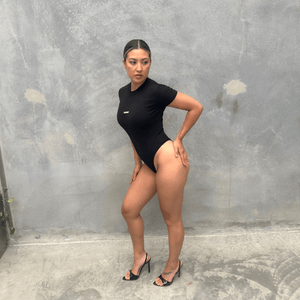 Woman in a black short sleeve bodysuit against a grey concrete wall posing from the side