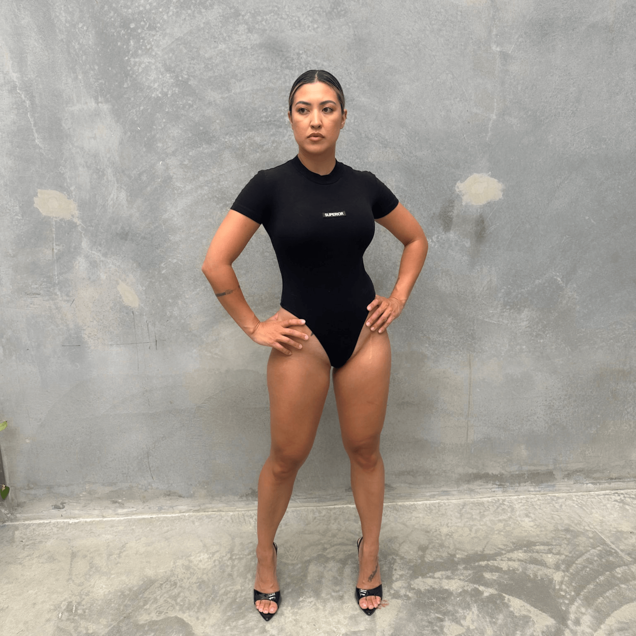 Woman wearing a black T-shirt short sleeve bodysuit standing against a grey concrete wall front of bodysuit 