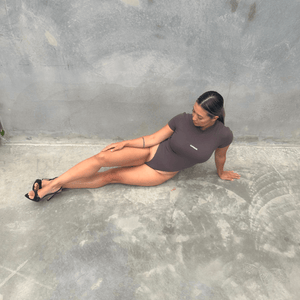 Woman in a charcoal short sleeve bodysuit laying on the floor against a grey wall front on 