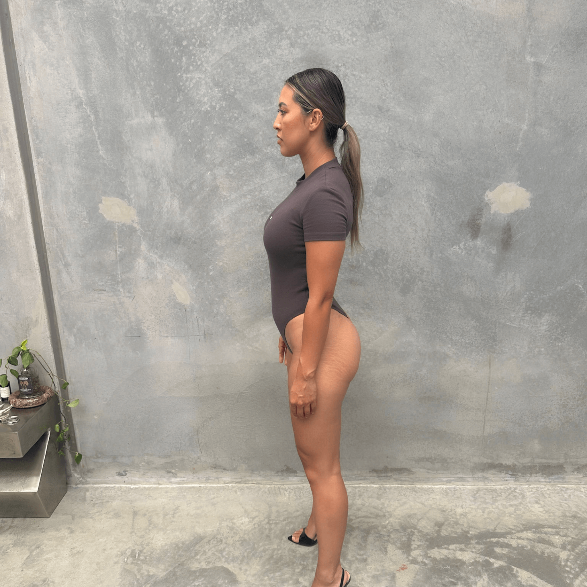 Woman in a charcoal short sleeve bodysuit standing against concrete wall side profile 