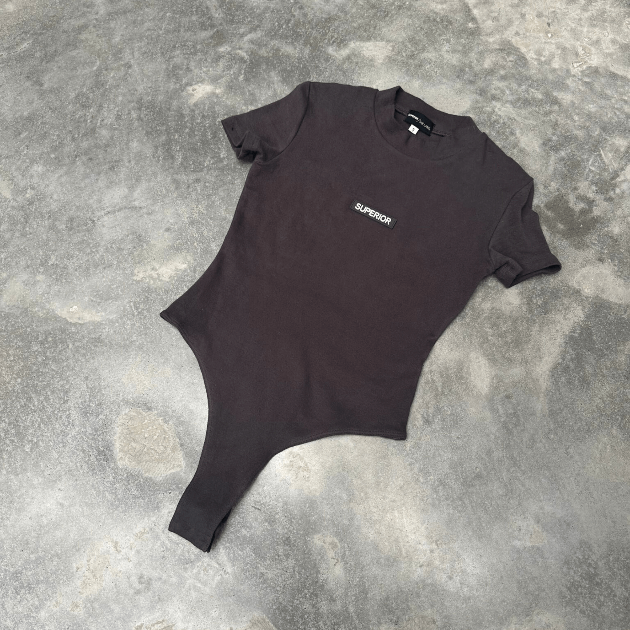 Black t-shirt short sleeve bodysuit with 'SUPERIOR' logo on a textured gray surface front on in flat lay position 