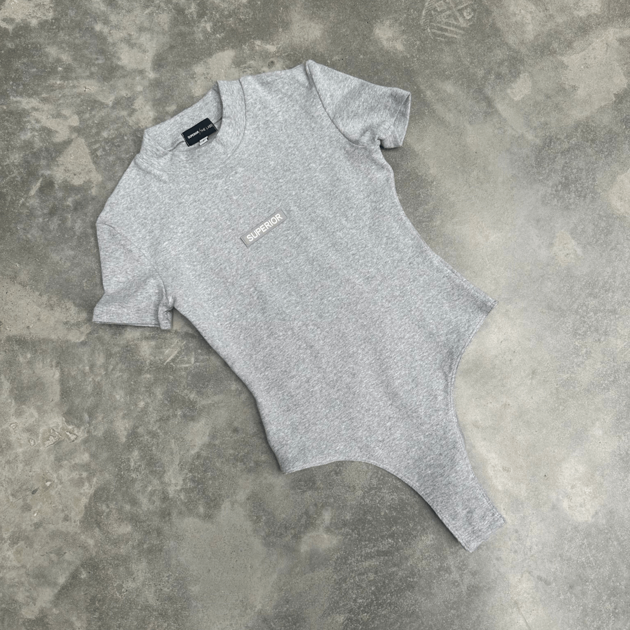 Grey t-shirt bodysuit with a small logo on a textured grey surface for flatlay